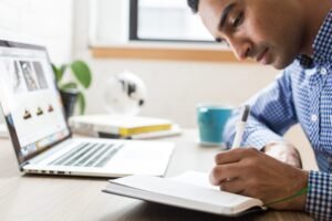 Man writing in a notebook while sitting at a desk with a laptop