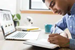 Man writing in a notebook while sitting at a desk with a laptop